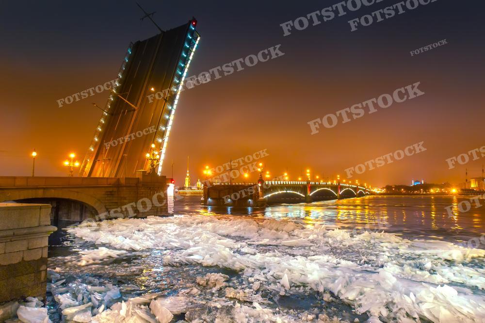 На фотографии представлен разведенный мост в ночном Санкт-Петербурге. Тёплый свет фонарей окрашивает ночной город в золотистые тона, и на заднем плане виднеется силуэт Петропавловской крепости. Эта фотография идеально подойдёт для туристических проспектов, публикаций о ночной жизни Петербурга или книг о русской культуре и истории.